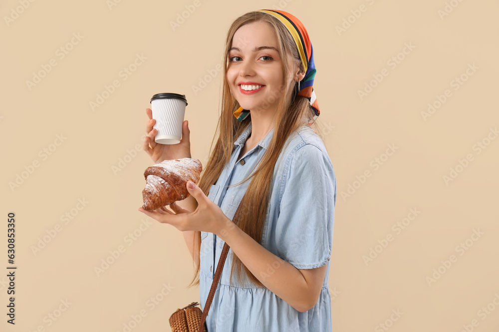 Young woman with tasty croissant and cup of coffee on beige background