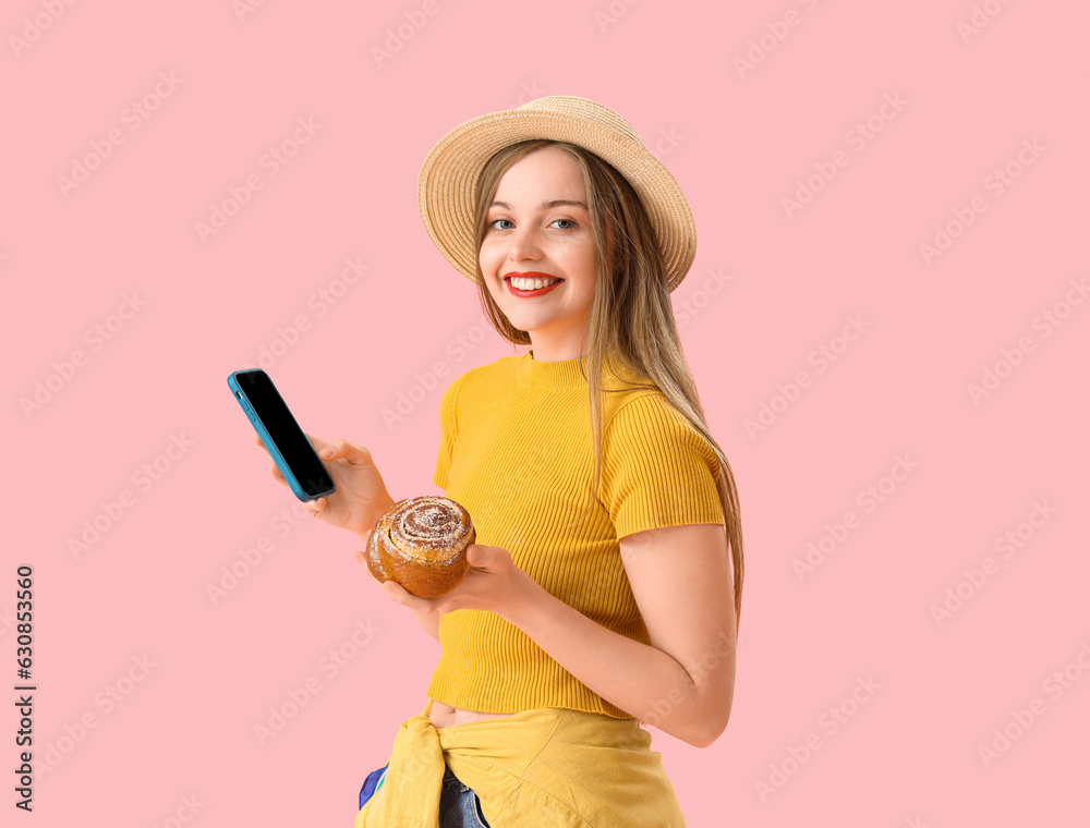 Young woman in hat with tasty bun and mobile phone on pink background