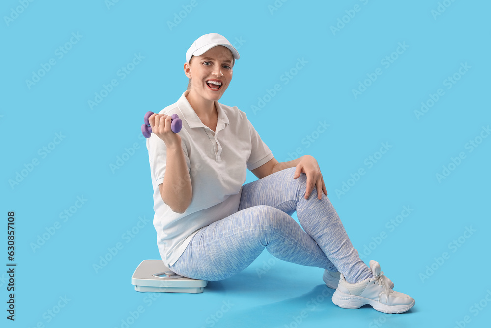 Young overweight woman with dumbbells and scales sitting on blue background