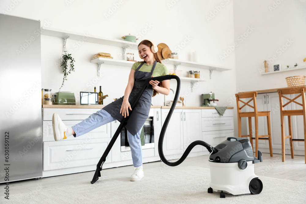 Young woman with headphones vacuuming carpet in kitchen