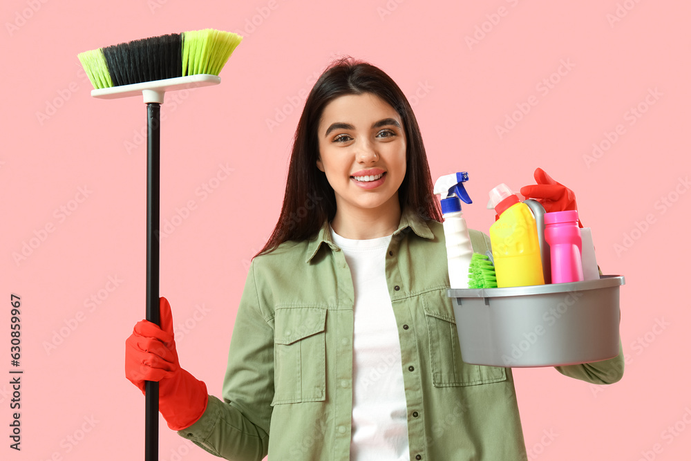 Young woman with broom and bucket of cleaning supplies on pink background