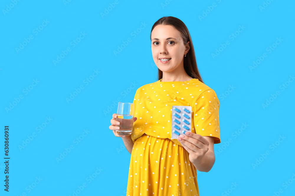 Young pregnant woman with glass of water and Folic Acid pills on blue background