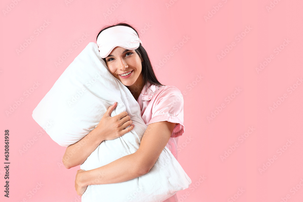 Young woman with sleeping mask hugging pillow on pink background