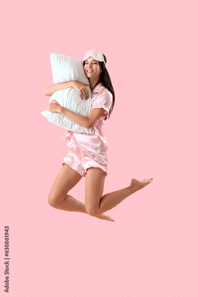 Young woman in pajamas with pillow jumping on pink background