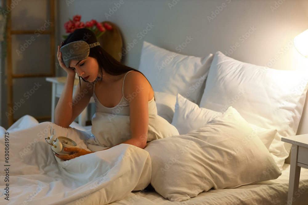 Upset young woman with alarm clock in bedroom at night