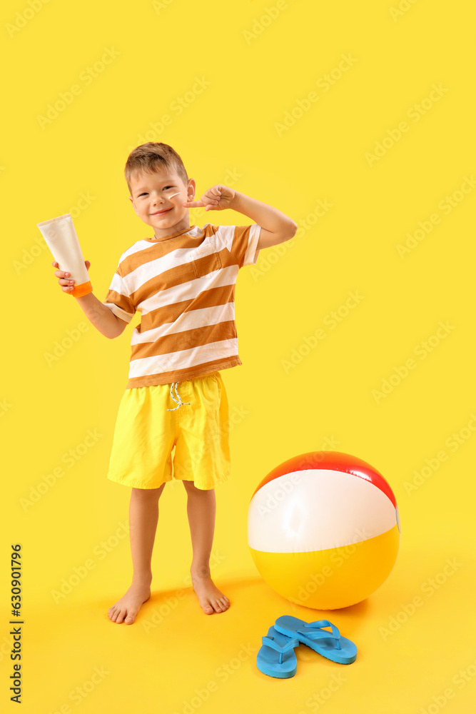 Little boy with bottle of sunscreen cream and inflatable ball on yellow background