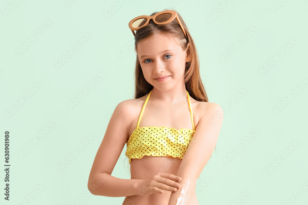 Little girl with sunglasses applying sunscreen cream on pale green background