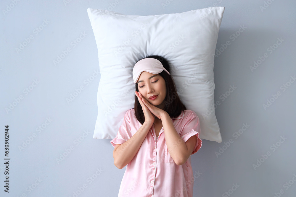 Beautiful Asian woman in pajamas with pillow on grey background