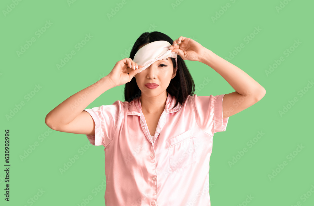 Beautiful Asian woman in pajamas with sleeping mask on green background