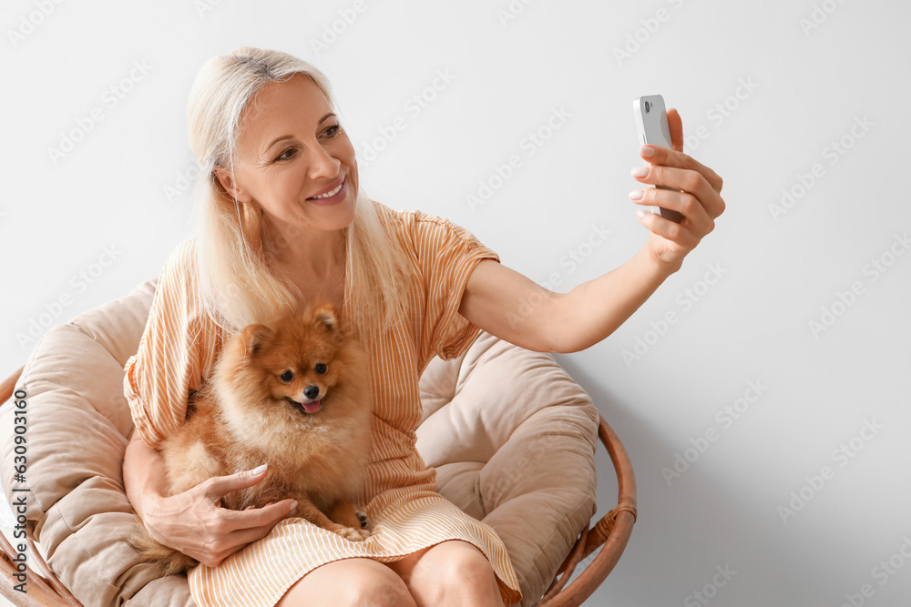 Mature woman with Pomeranian dog taking selfie in armchair near light wall