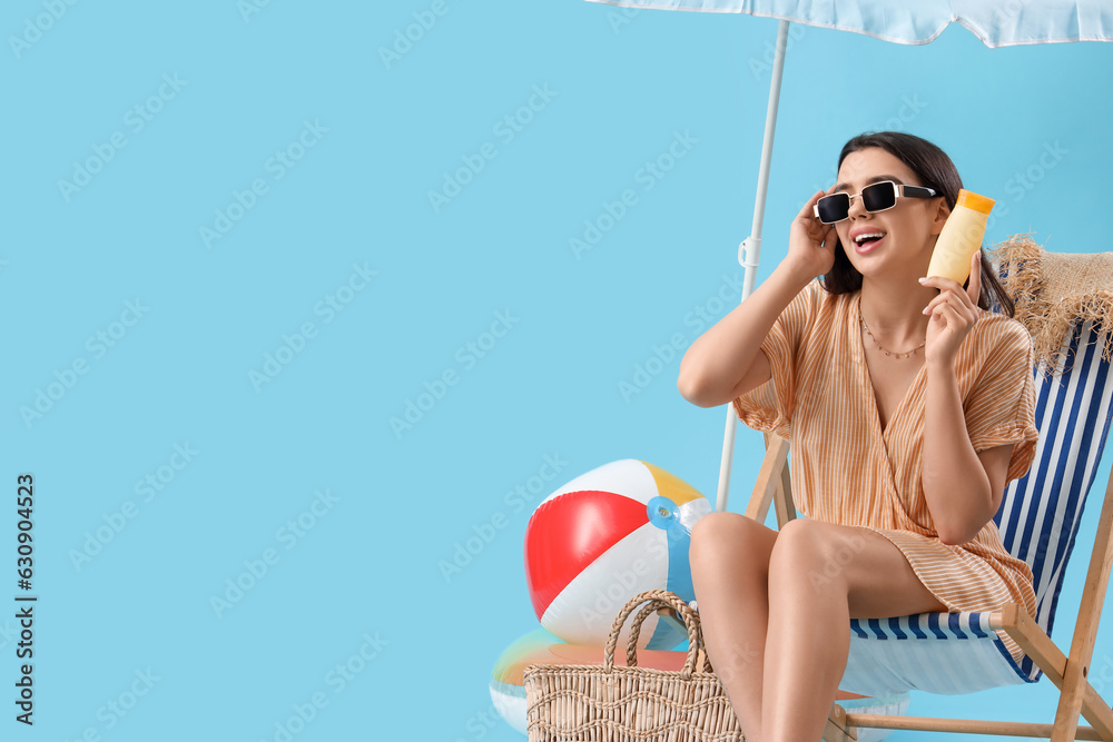 Young woman with sunscreen cream in deck chair on blue background