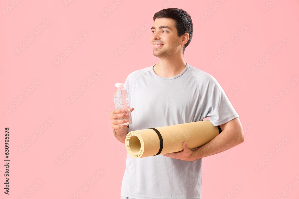 Sporty young man with bottle of water and fitness mat on pink background