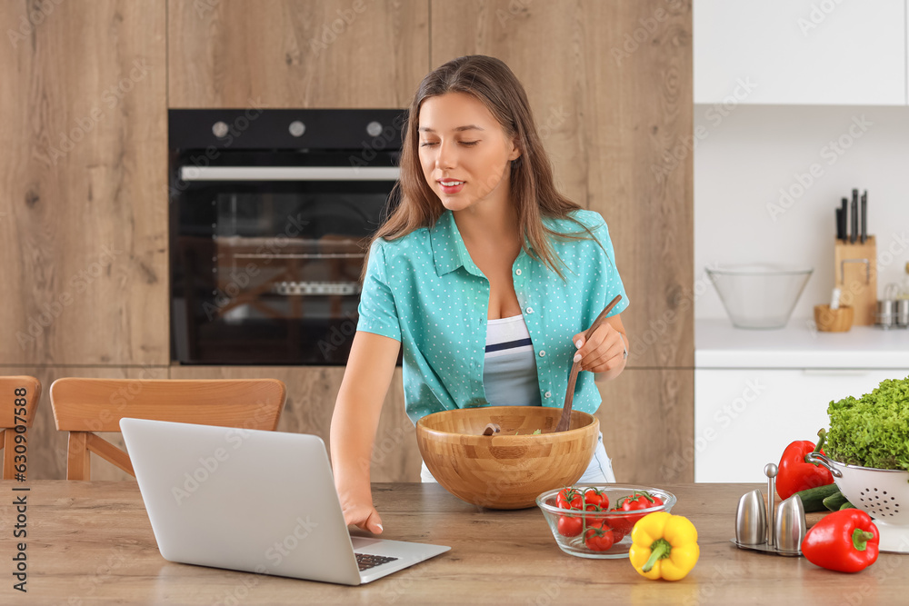 Young woman with laptop preparing vegetable salad in kitchen