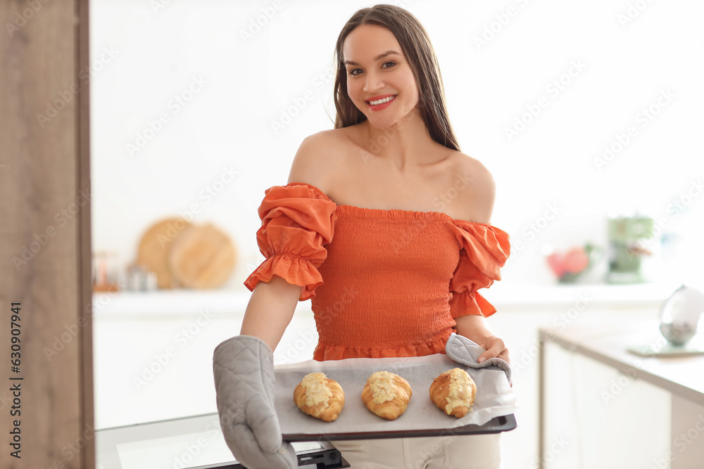 Young woman holding baking tray with croissants in kitchen