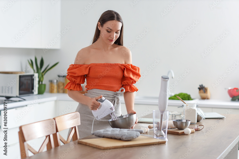 Young woman whipping eggs with mixer for dough in kitchen