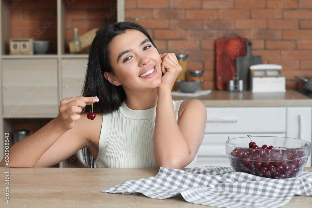 Beautiful young woman with bowl of ripe cherries sitting at table in kitchen