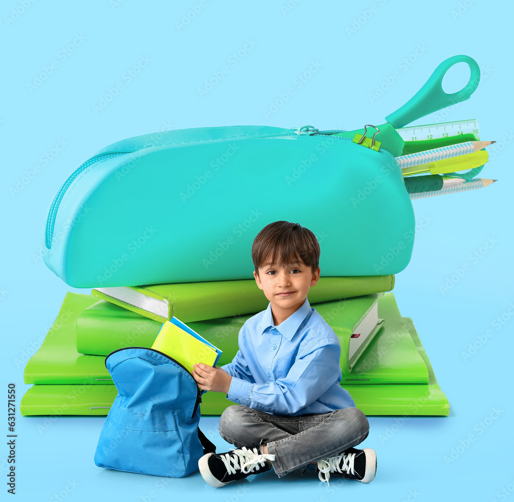 Little schoolboy putting notebooks in backpack on color background