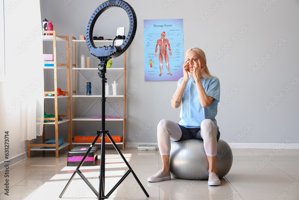 Mature physiotherapist doing face building exercise while recording video in rehabilitation center