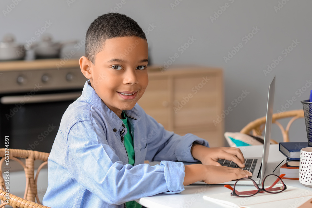 Little African-American boy with laptop studying computer sciences online in kitchen