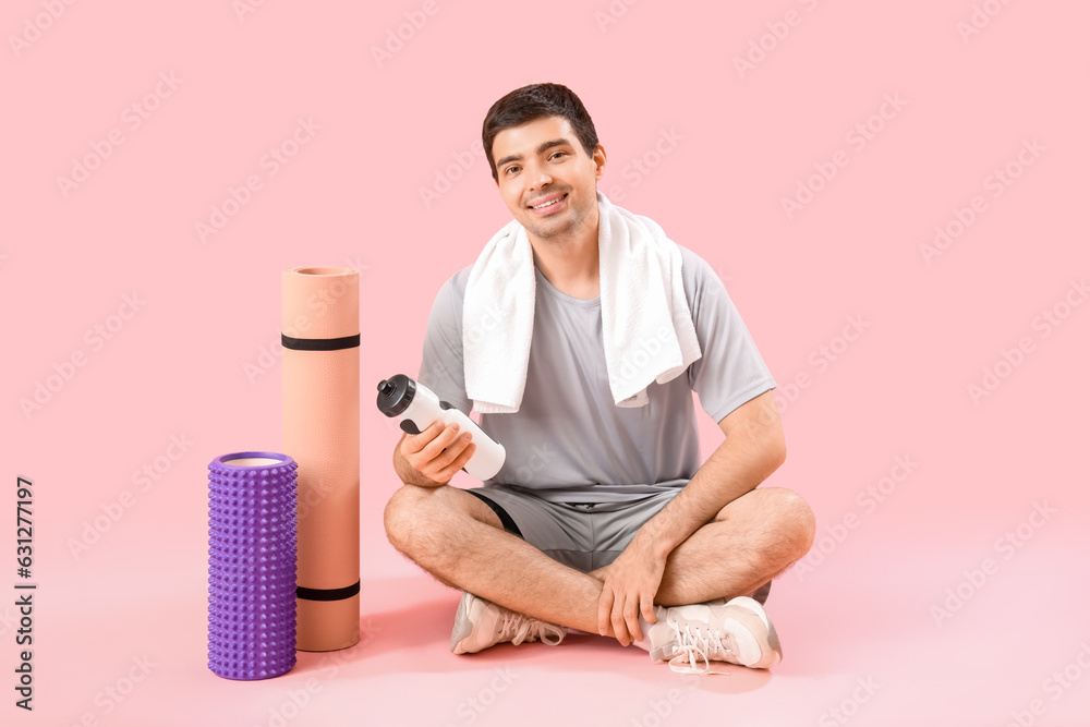 Sporty young man with bottle of water and fitness mats sitting on pink background