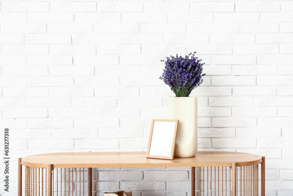 Vase with beautiful lavender flowers and blank frame on shelving unit near light brick wall in room