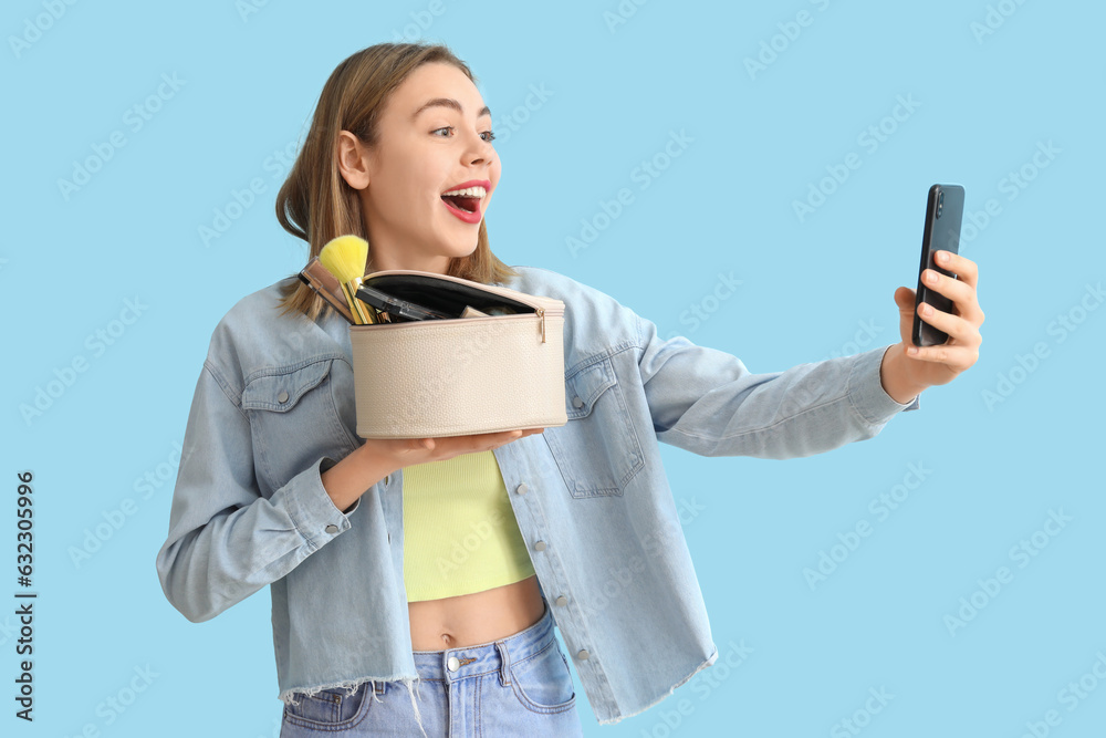 Young woman with cosmetic bag and mobile phone taking selfie on blue background