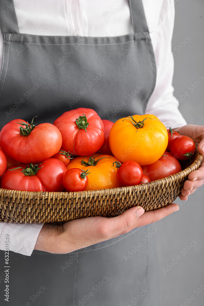 Woman holding wicker bowl with different fresh tomatoes on grey background, closeup