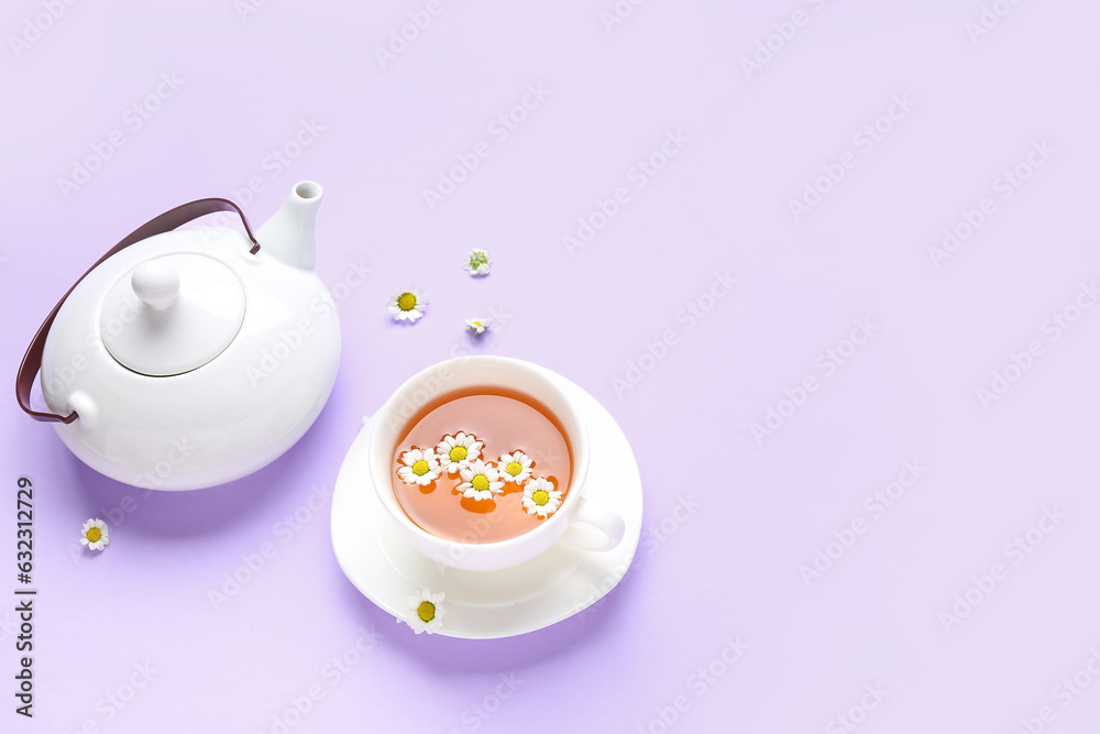 Teapot with cup of natural chamomile tea and flowers on lilac background