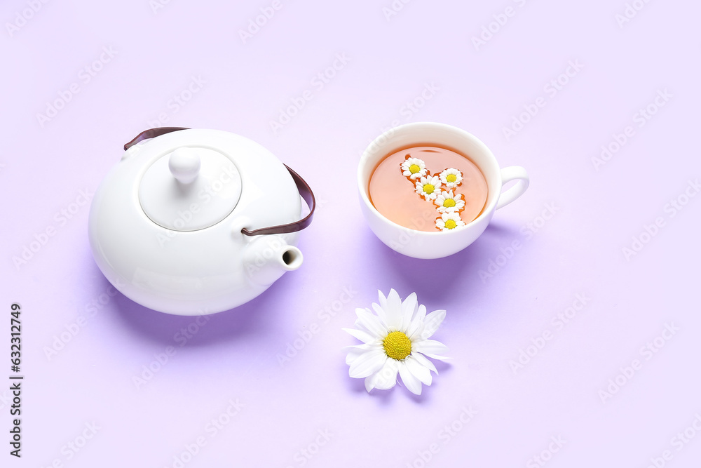 Teapot with cup of natural chamomile tea and flowers on lilac background