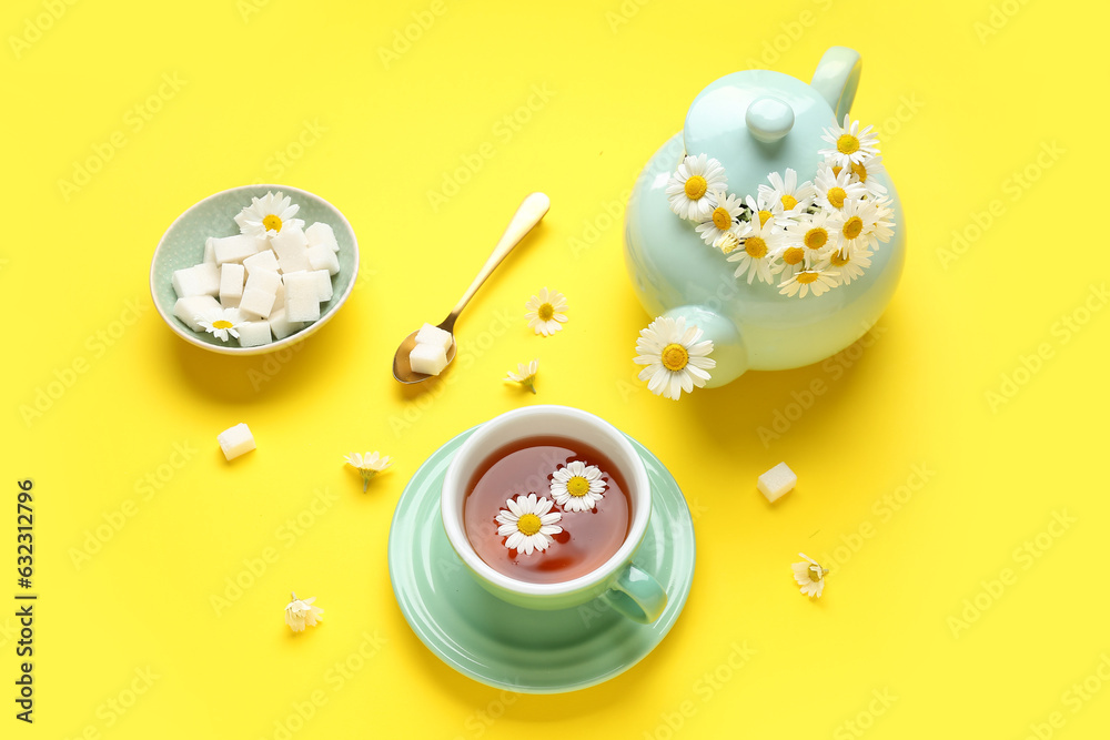 Teapot with cup of natural chamomile tea, sugar cubes and flowers on yellow background