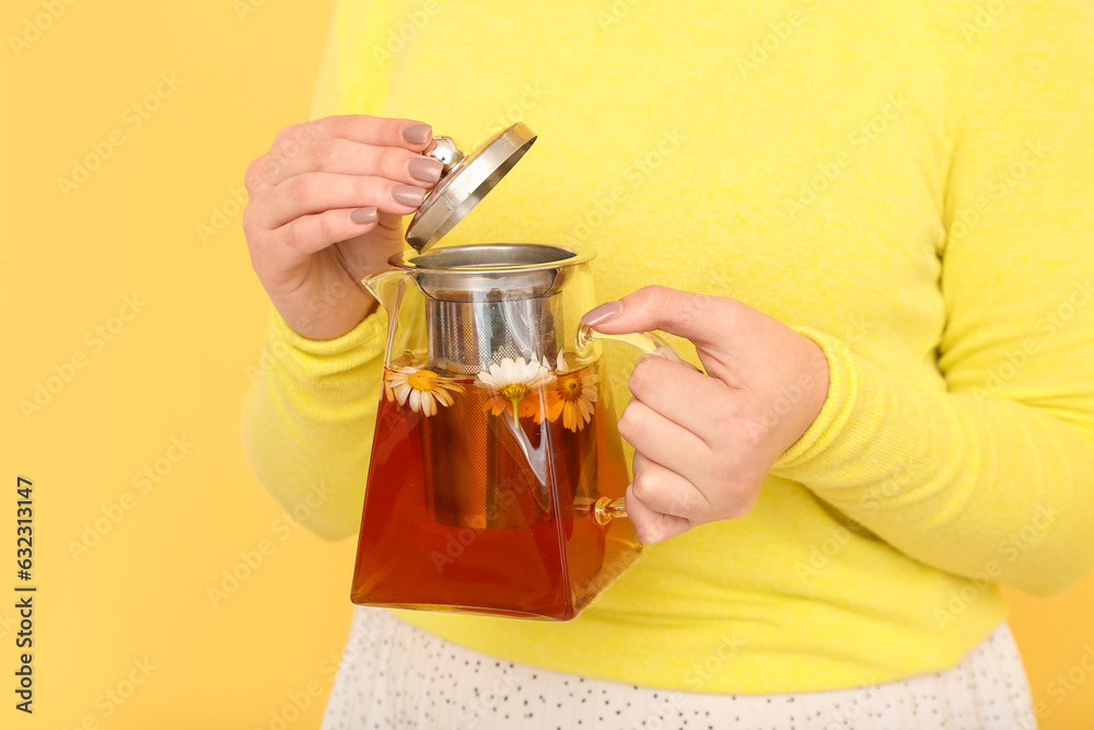 Beautiful young woman with teapot of chamomile tea near yellow wall