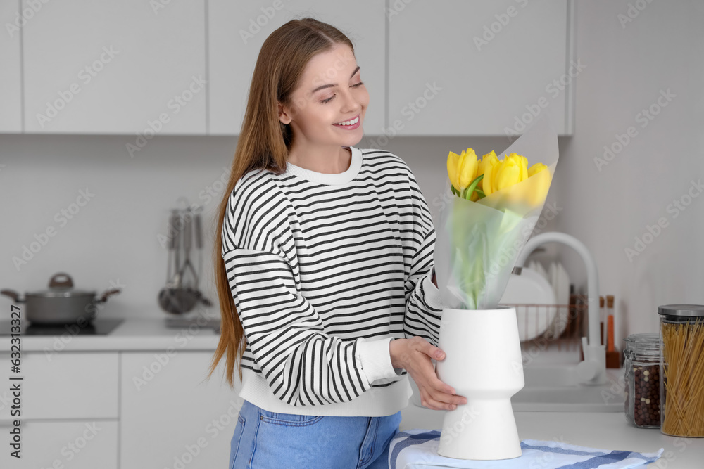 Beautiful young woman putting bouquet of yellow tulip flowers into vase in modern kitchen