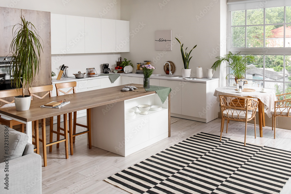 Interior of modern kitchen with white counters, island and dining table near big window