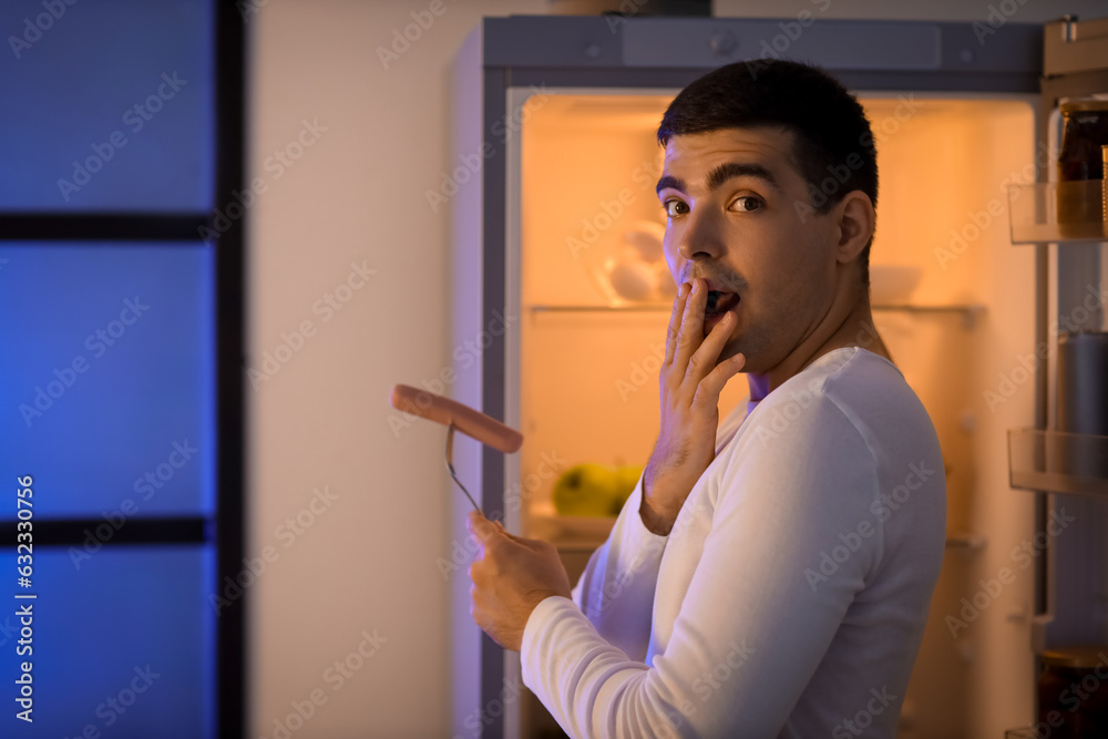 Scared young man with sausage near open fridge in kitchen at night