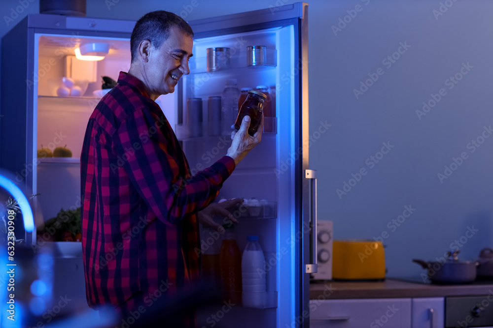 Hungry mature man with jam near open fridge in kitchen at night