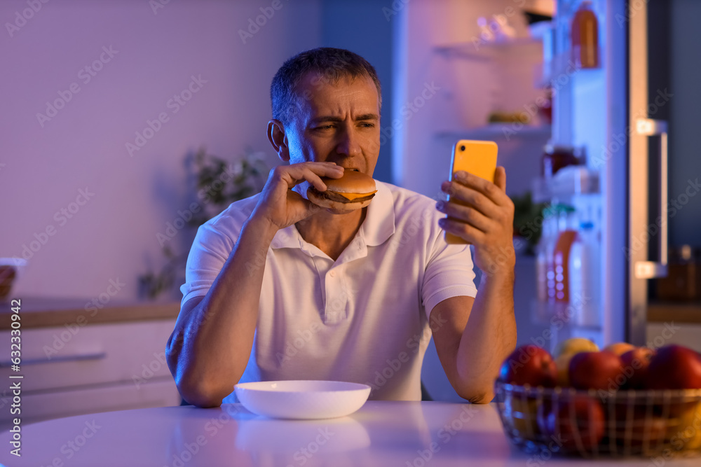 Hungry mature man with burger and mobile phone in kitchen at night