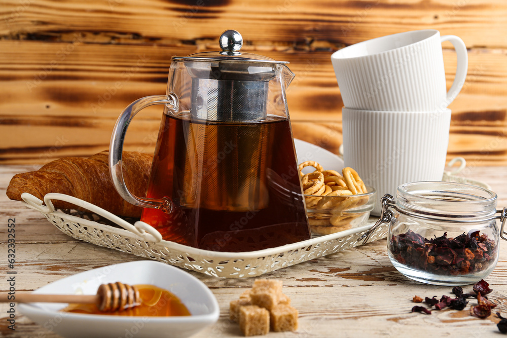 Glass teapot with tea, cups, croissant and pretzels on light wooden table