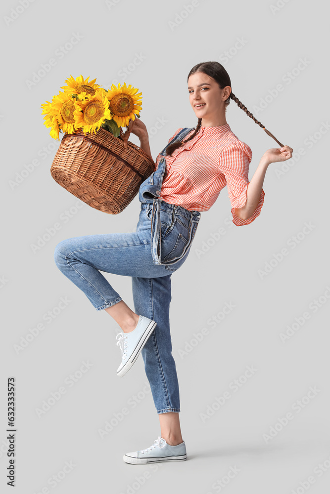 Happy young woman with wicker basket full of beautiful sunflowers on grey background