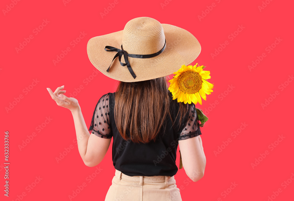 Young woman in wicker hat with beautiful sunflower on red background, back view