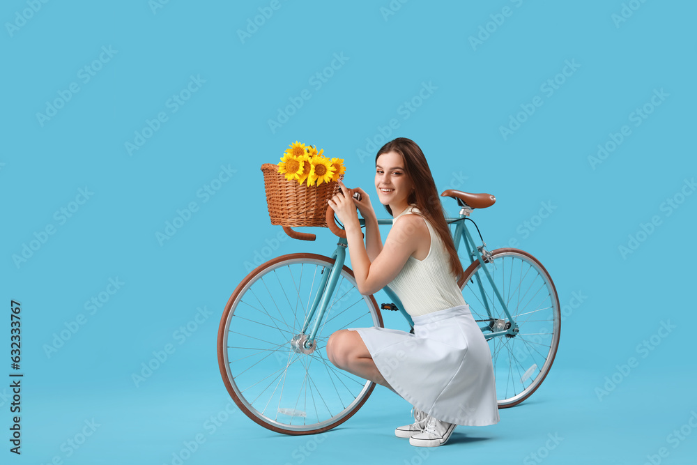 Young happy woman with bicycle and wicker basket full of beautiful sunflowers on blue background