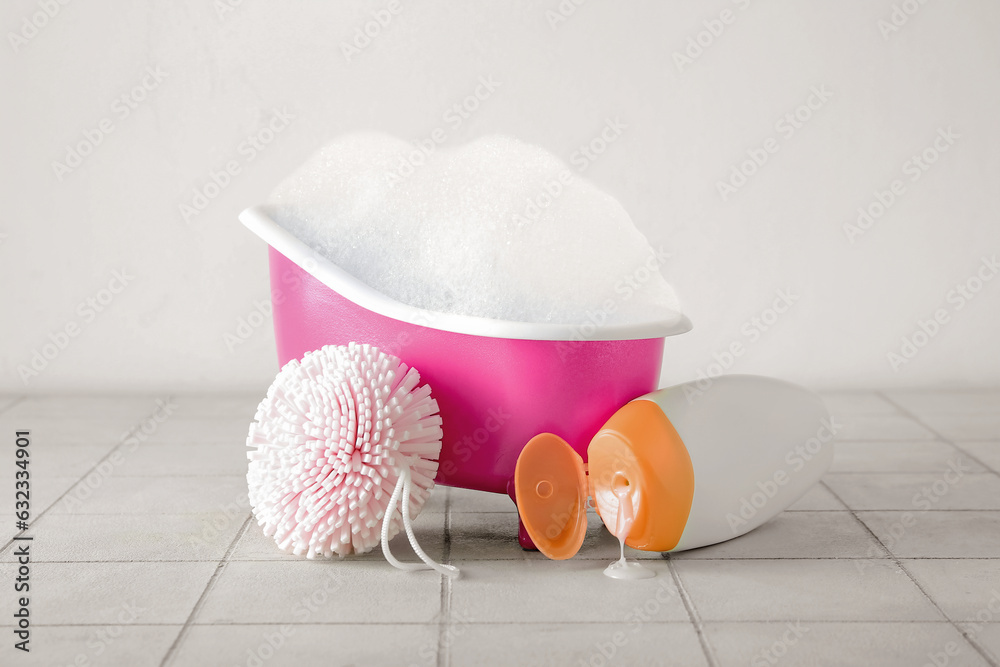 Small bathtub with foam, sponge and bottle of cosmetic product on light tile background