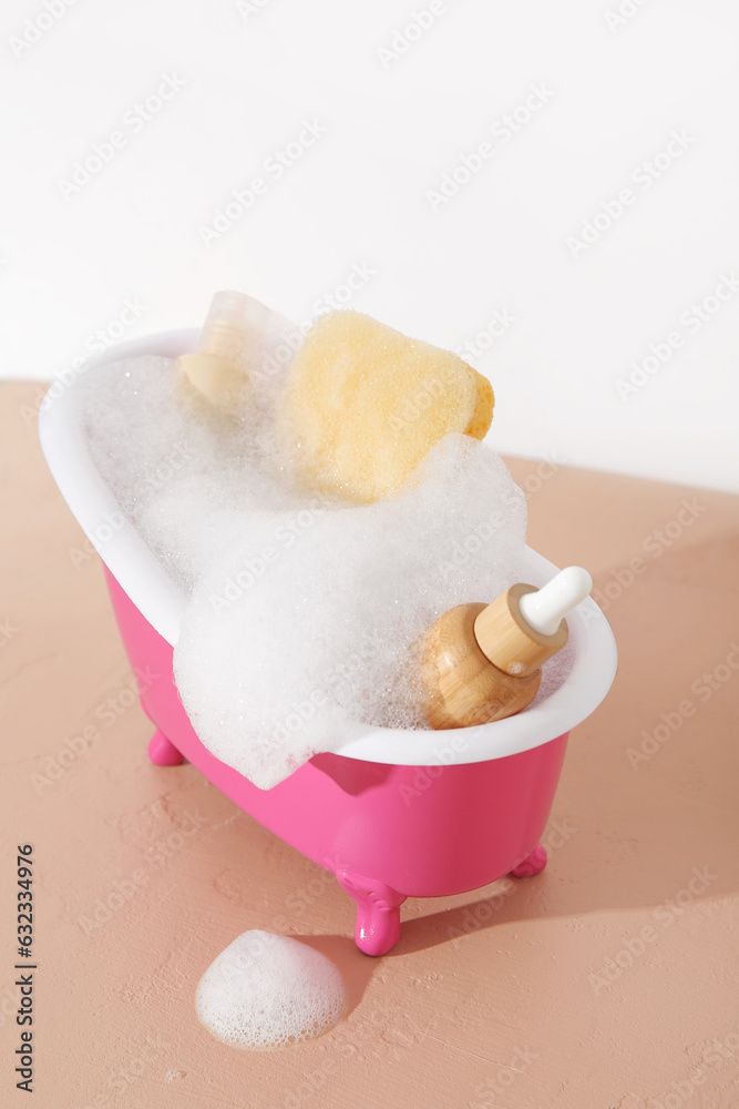 Small bathtub with foam, cosmetic products and sponge on color table against light background