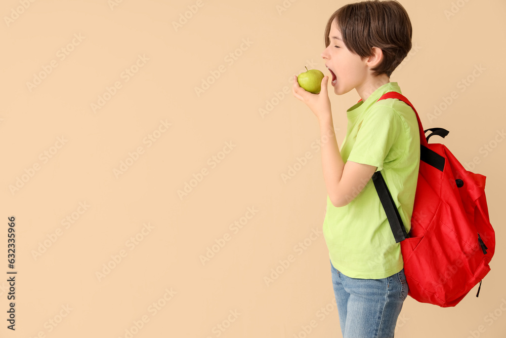 Little boy with backpack eating fresh apple on beige background