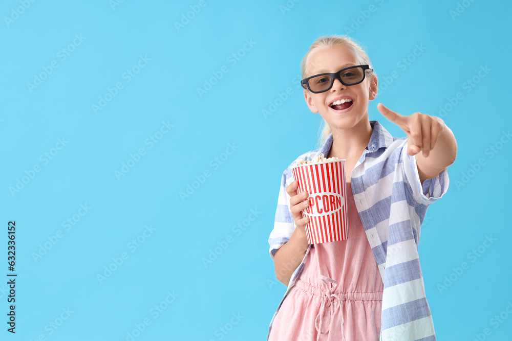 Happy girl in 3D cinema glasses with bucket of popcorn pointing at something on blue background