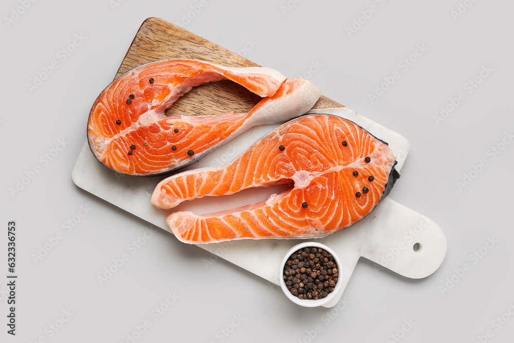 Board with raw salmon steaks and bowl of peppercorn on grey background