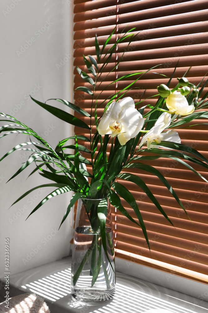 Vase with orchid flowers and tropical leaves on window sill