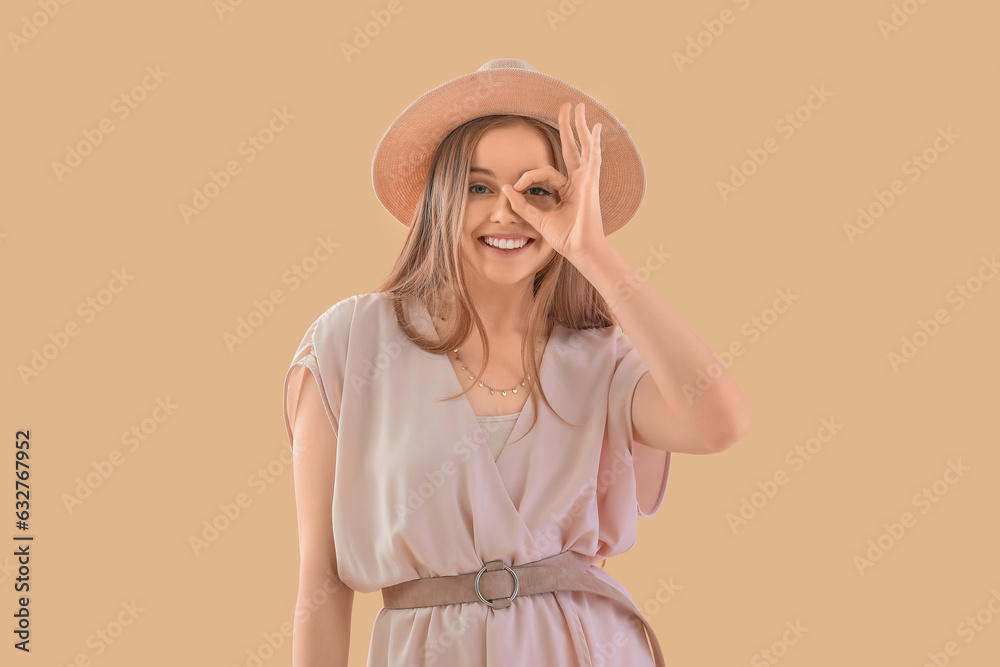 Young woman in hat showing OK on beige background