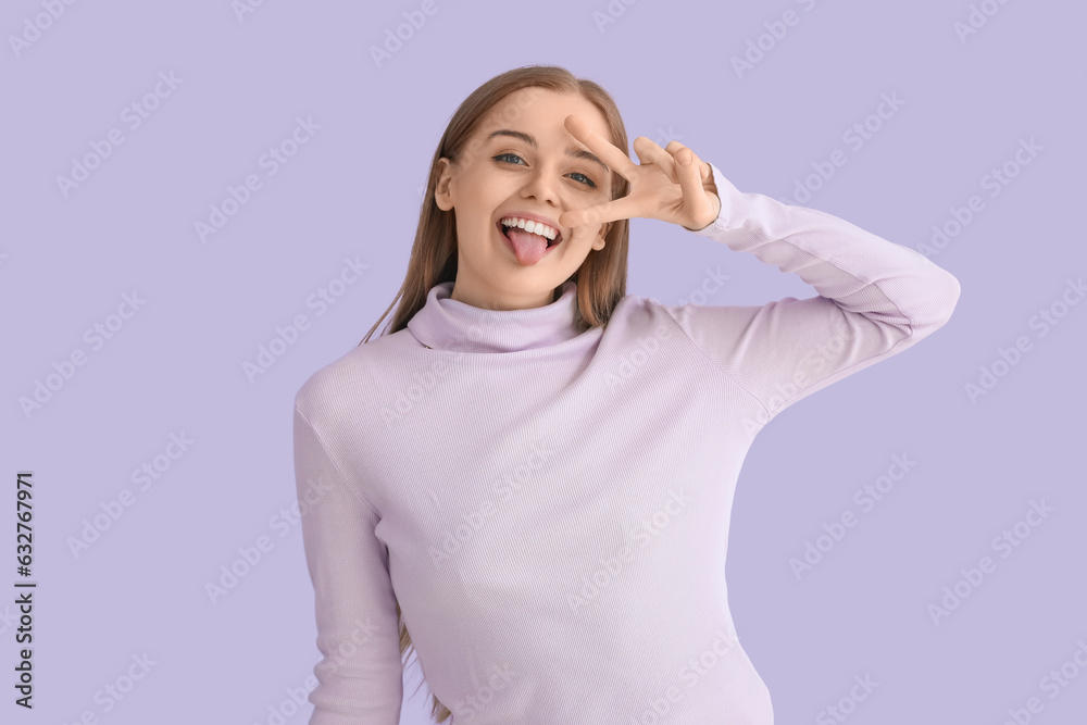 Funny young woman in sweater showing victory gesture on lilac background