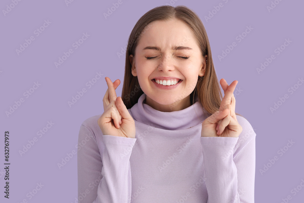 Young woman in sweater crossing fingers on lilac background, closeup