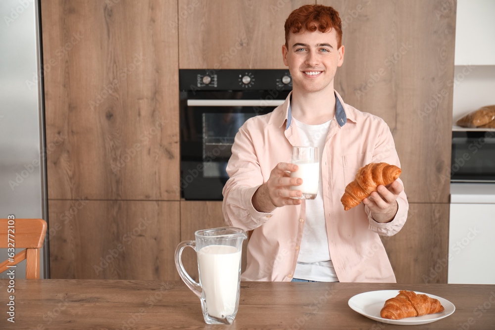 Young redhead man with glass of milk and croissant in kitchen